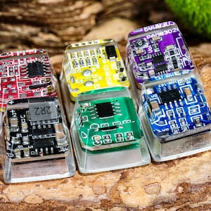 May include: Six colorful keycaps with circuit board designs. The keycaps are made of clear resin and feature different colored circuit boards. The keycaps are arranged in a row on a wooden surface.