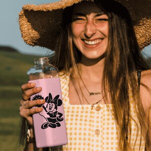May include: A pink reusable water bottle with a black and white graphic of Minnie Mouse. The bottle has a straw lid and is held by a person wearing a straw hat and a yellow and white gingham dress.