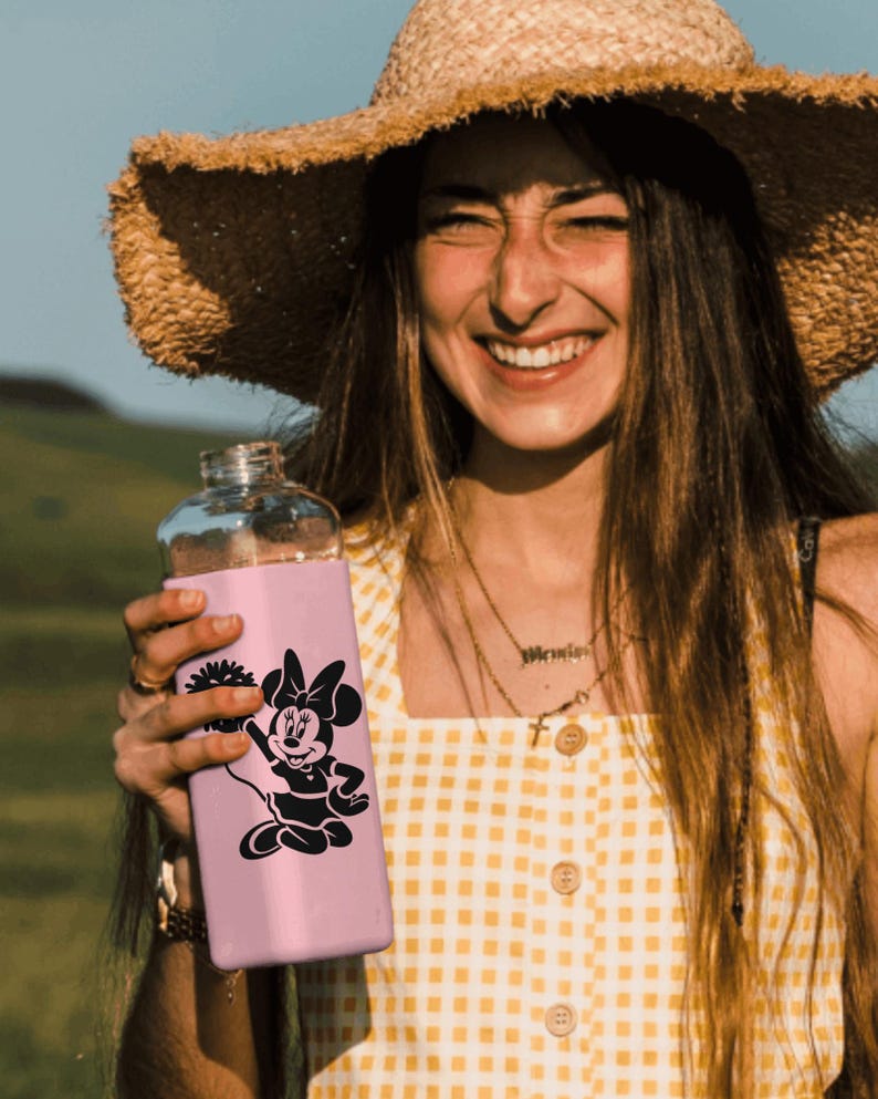 May include: A pink reusable water bottle with a black and white graphic of Minnie Mouse. The bottle has a straw lid and is held by a person wearing a straw hat and a yellow and white gingham dress.