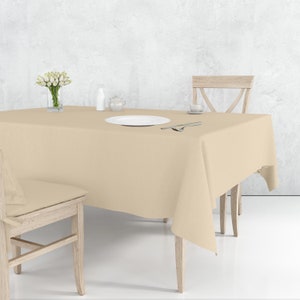 Può includere: Una tovaglia beige è stesa su un tavolo di legno con due sedie. Un piatto bianco con posate è sul tavolo, insieme a un vaso di fiori bianchi e una piccola teiera.