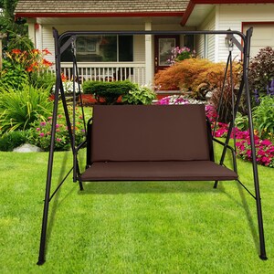 May include: A black metal swing set with a brown cushioned seat. The swing is in a grassy garden with flowers and a house in the background.