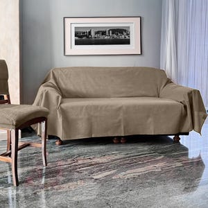 Può includere: Una fodera per divano color talpa drappeggiata su un divano a tre posti. La fodera è realizzata in un tessuto morbido e ha un design semplice ed elegante. Una stampa incorniciata in bianco e nero di un paesaggio urbano è appesa sopra il divano. Una sedia abbinata è a sinistra.