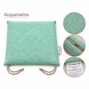 Peut inclure: Un coussin d'assise carré couleur aigue-marine avec une surface en tissu texturé, doté de deux attaches en cordon beige aux coins inférieurs. Le coussin comprend une fermeture éclair latérale et une étiquette blanche avec le texte Il Tuo Artigiano Design.