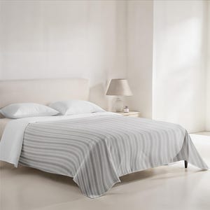 Puede incluir: Una cama blanca con una colcha a rayas grises y blancas. La cama tiene dos almohadas blancas y una cabecera blanca. Una lámpara está sobre una mesita de noche junto a la cama.