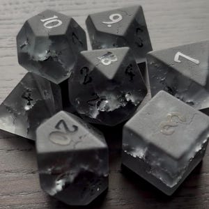 May include: A set of seven gray, opaque dice with white numbers. The dice have a textured, cracked surface. The numbers on the dice are 10, 9, 8, 7, 6, 5, and 4.