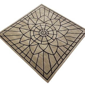 May include: A square beige rug with a black geometric design. The design features a large circular pattern in the center with radiating lines.