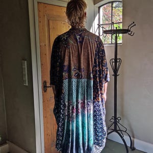 May include: A long, patchwork-style robe with a mix of patterns and colors, including brown, blue, and teal. The robe has a loose fit and appears to be made of a soft fabric. The garment is worn over blue jeans.