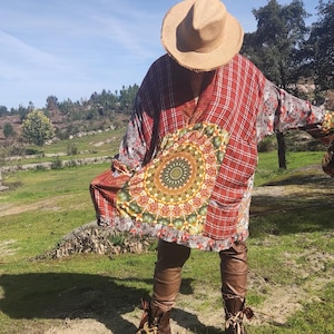 Op de afbeelding: Een persoon draagt een rood geruite kimono met een groot mandala-ontwerp op de voorkant. De kimono heeft lange mouwen en wordt gecombineerd met een bruine leren broek en laarzen. Er wordt een strohoed gedragen.