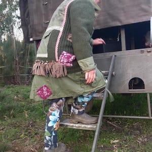 Könnte beinhalten: Eine Person in einer Patchwork-grünen Jacke, einem olivgrünen Rock und einer Leggings mit Blumenmuster steigt eine Metallleiter hinauf. Die Jacke hat rosa Spitzen- und Fransen Details. Die Person trägt braune Stiefel.