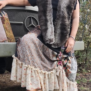 Könnte beinhalten: Eine Frau trägt ein schwarzes Oberteil und eine Weste aus Kunstfell, kombiniert mit einem Patchwork-Rock und braunen Cowboy-Stiefeln. Der Rock hat florale und gemusterte Stoffe mit einem Rüschensaum. Der Hintergrund zeigt ein Militärfahrzeug.