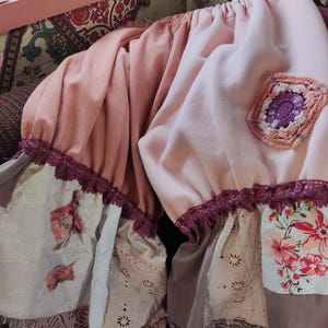 Può includere: Due gonne a strati nei toni del rosa e del beige, con pannelli in pizzo e floreali. È visibile una toppa quadrata all'uncinetto. I bordi sono rifiniti con pizzo viola scuro. Le gonne sono arruffate.