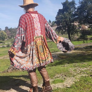 Op de afbeelding: Een patchworkjas met een rood ruitpatroon, bloemen- en paisleyprints en een gefranjerde zoom. De jas wordt gedragen met een bruine leren broek, bruine laarzen en een strohoed. Een patroonemmerhoed wordt in de hand gehouden.