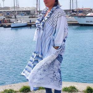 Pode incluir: Uma mulher vestindo uma roupa longa e fluida, branca e azul com um desenho de mapa. A roupa tem um padrão listrado nas mangas e uma gola com babados. Ela está usando uma faixa preta, jeans azuis e sandálias. O fundo apresenta barcos e água.