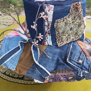 Op de afbeelding: Een patchwork hoed met een denim basis en diverse stoffen patches in tinten blauw, bruin en bloemenprints. De hoed is versierd met een takje en kleine roze bloemaccenten, wat een uniek, handgemaakt accessoire creëert.