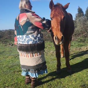 Op de afbeelding: Een persoon in een patchworkjas en rok staat naast een bruin paard in een grasveld onder een blauwe lucht. De jas heeft diverse patronen en kleuren, terwijl de rok meerdere lagen en texturen heeft.