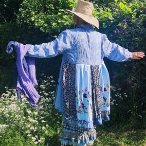 Op de afbeelding: Een lichtblauwe tuniek met lange mouwen, witte bloemenborduursels op de bovenrug, een patchwork rokgedeelte met bloemen- en paisley-patronen en een gelaagde ruchezoom. Een paarse sjaal wordt vastgehouden en een strooien hoed wordt gedragen.