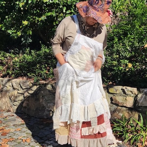 Puede incluir: Una persona con un sombrero marrón con estampado floral, una camisa color canela y un delantal blanco con detalles de encaje y patchwork. El delantal está superpuesto a una falda con patchwork y encaje similares. Botas marrones completan el atuendo.
