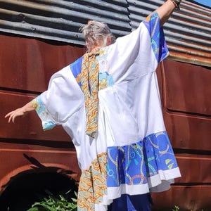 Op de afbeelding: Een witte, blauwe en oranje kimono-achtige top met een bijpassende sjaal en een blauwe rok met ruches. De kimono heeft blauwe en oranje patronen op de mouwen en zoom. De rok is effen blauw.
