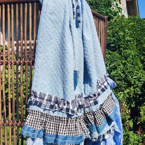 May include: A light blue textured fabric skirt featuring a drawstring waist and multiple layered ruffles at the hem. The ruffles consist of alternating bands of blue and white gingham, black and white checkered fabric, and denim-like material.