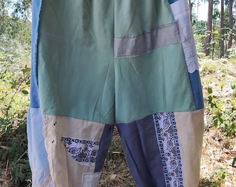 Op de afbeelding: Patchworkbroek in tinten groen, blauw en beige hangt aan een houten hanger. De broek heeft een verscheidenheid aan stoffen, waaronder denim, kant en katoen met patronen. De manchetten zijn gerimpeld en de taille is elastisch.