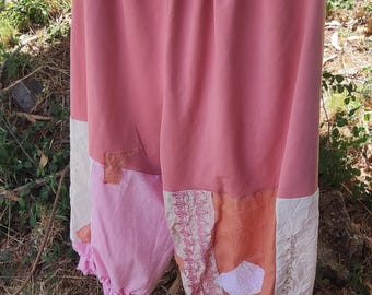 Op de afbeelding: Een roze en perzikkleurige broek met een geplooide tailleband en gerimpelde manchetten hangt aan een houten kleerhanger. De broek heeft patchworkdetails in verschillende tinten roze, perzik en crème, met geborduurde accenten.