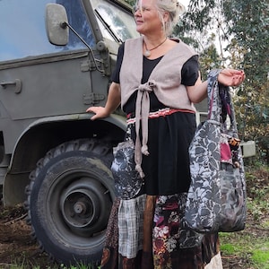 Op de afbeelding: Een vrouw draagt een zwarte jurk, een beige vest en een patchwork rok, met een grote draagtas met bloemenpatroon en houten handvat. De rok heeft diverse stofpatronen. Ze staat bij een militair voertuig.