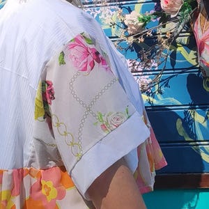 Peut inclure: Haut de style kimono blanc avec des manches à imprimé floral et un ourlet à motifs colorés. Les manches présentent des roses roses et un motif de chaîne sur un fond clair. L'ourlet présente des motifs floraux orange, roses et jaunes.