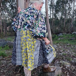 Pode incluir: Uma mulher vestindo um vestido longo de patchwork multicolorido com um top de estampa floral. O vestido tem um padrão xadrez preto, branco e amarelo, uma estampa de leopardo e uma estampa floral. Ela está usando botas marrons e está em uma floresta.