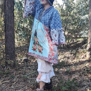 Può includere: Giacca stile kimono blu fantasia con maniche ampie, abbinata a una tunica azzurra con fantasia floreale. L'outfit comprende un cappello nero, collane a strati e stivali marroni. L'ensemble è indossato all'aperto in un'area boschiva.