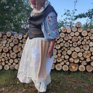 Puede incluir: Un atuendo en capas con una blusa azul con estampado floral, un chaleco negro y una falda de encaje blanca. Botas de vaquero marrones con diseños rojos y azules completan el look. El fondo presenta una pila de troncos cortados y un cielo azul.