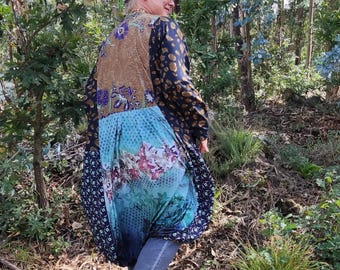 Może przedstawiać: Długa szata w stylu kimono z wzorem patchwork w odcieniach brązu, błękitu i kwiatowych nadruków. Szata jest noszona z niebieskimi dżinsami i beżowymi sandałami. Osoba spaceruje w leśnym otoczeniu.