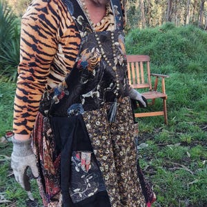 Könnte beinhalten: Eine Frau trägt einen braunen Hut, ein Tiger-Shirt und ein langes, schwarzes Patchwork-Kleid mit rotem Saum. Das Kleid hat mehrere Taschen und ein Blumenmuster. Sie steht in einem grasbewachsenen Bereich mit einem Holzstuhl im Hintergrund.