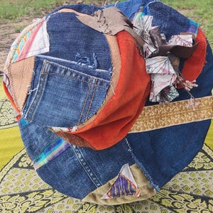 Op de afbeelding: Een ronde patchwork hoed gemaakt van gerecycled denim, oranje stof en andere textielsoorten. De hoed heeft een verscheidenheid aan texturen en kleuren, waaronder blauwe denim, oranje, beige en bloemmotieven. De hoed heeft een rand.