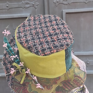 May include: A unique, patchwork hat with a tweed top in black, brown, and orange. The hat features a green brim, paisley patterned fabric, and is adorned with a feather and pink floral accents. A stylish accessory.