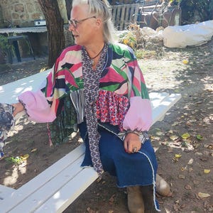 Peut inclure: Une veste en patchwork composée de panneaux à motifs floraux, abstraits et unis roses avec des manches longues, associée à une jupe en jean bleu foncé dotée d'un ourlet effiloché. La veste comprend une longue écharpe fine à motif noir et blanc.