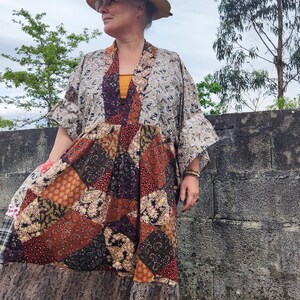 Pode incluir: Uma mulher vestindo um quimono estampado em marrom e branco com uma saia de patchwork. Ela está usando um chapéu marrom e sandálias verdes. A saia é feita de diferentes tecidos com padrões florais e paisley.