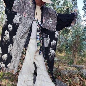 May include: A patchwork kimono-style jacket with black, gray, and floral patterned panels. The jacket is worn with white pants and a straw hat. The outfit is accessorized with a patterned scarf and a beaded necklace. The sandals have a brown sole and white straps.