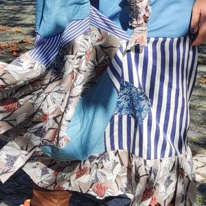 Puede incluir: Una falda patchwork con telas azules, blancas y estampadas con flores. La falda presenta rayas verticales, volantes y un panel azul. Botas marrones con detalles florales completan el look.