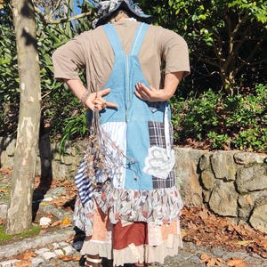May include: A patchwork denim apron with a variety of fabric patterns, including floral, plaid, and lace. The apron has a ruffled hem and is worn with brown boots. A bundle of dried flowers is held in front.