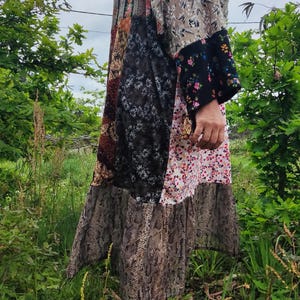 Könnte beinhalten: Ein langes, mehrfarbiges Patchworkkleid mit Blumenmuster und einem Schlangenhautmuster am Saum. Das Kleid hat einen Rüschen-Saum und wird von einer Person getragen, die in einem grasbewachsenen Bereich steht.