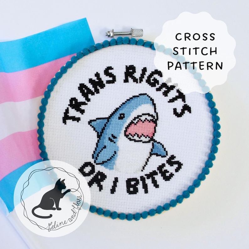 A cross stitch of a blue shark with an open mouth and its teeth showing, with the wording ‘trans rights or I bites’ in a circle around it. The design has been framed in a circular embroidery hoop with blue pom-pom trim and a trans flag is behind the hoop.