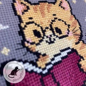 A close up of a cross stitch of a cute ginger cat reading a book