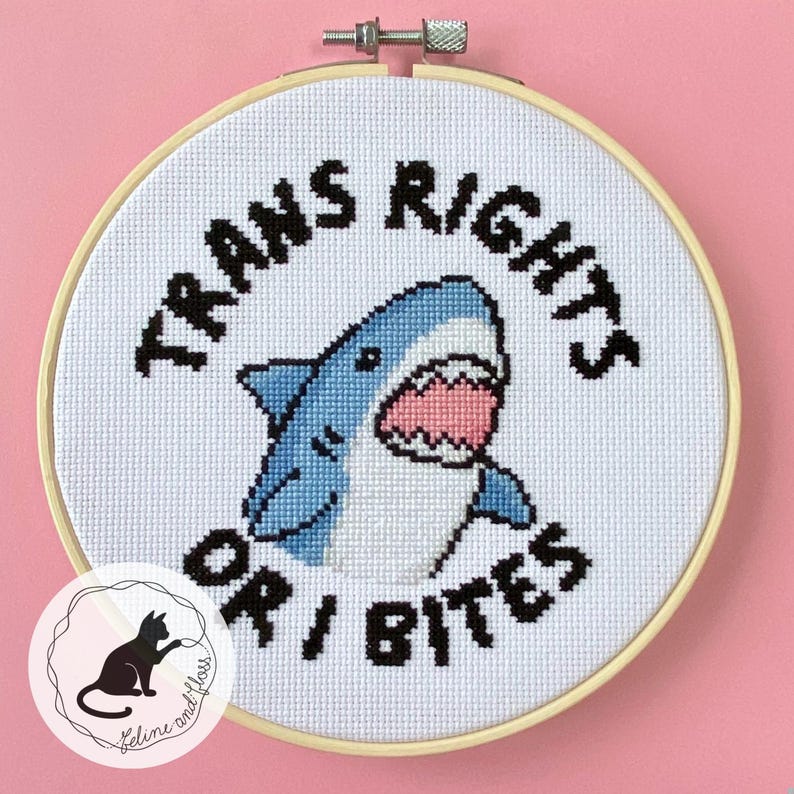 A cross stitch of a blue shark with an open mouth and its teeth showing, with the wording ‘trans rights or I bites’ in a circle around it.