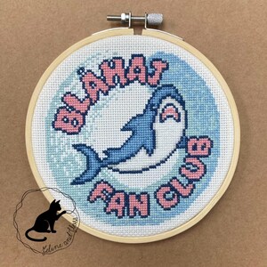 A blue and white cross-stitch with a blue shark and the text "BLAHAJ FAN CLUB" in pink letters.