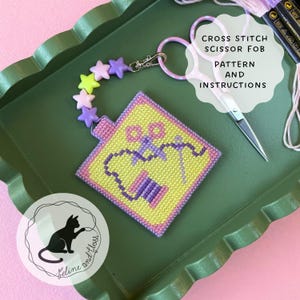 Puede incluir: Un llavero de tijeras de punto de cruz con fondo amarillo y borde rosa. El llavero presenta un diseño de tijeras e hilo. El texto "CROSS STITCH SCISSOR FOB PATTERN AND INSTRUCTIONS" es visible. Se adjunta una cadena de cuentas en forma de estrella.