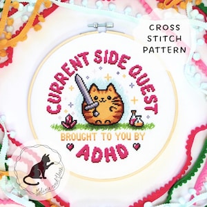 Peut inclure: Un motif de point de croix dans un cercle en bois. Il présente un chat de dessin animé avec une épée, avec le texte "CURRENT SIDE QUEST BROUGHT TO YOU BY ADHD." Comprend un cristal rose, une potion et "CROSS STITCH PATTERN."