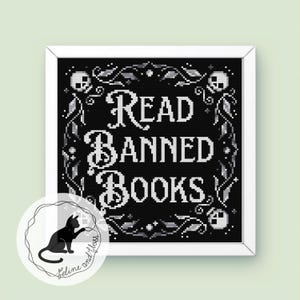 May include: A framed cross-stitch with the text "Read Banned Books" in white on a black backdrop. The design features skulls and floral motifs. A circular logo with a black cat is in the lower left corner.