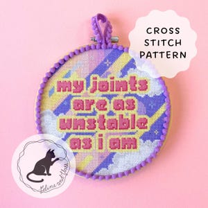 Pode incluir: Um padrão de ponto cruz em um bastidor de madeira com o texto "my joints are as unstable as i am" em letras rosa. O fundo apresenta um design colorido com tons de roxo, amarelo e azul. Um logotipo de gato está no canto inferior esquerdo.
