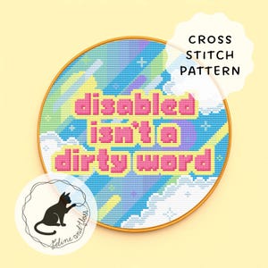 May include: A cross-stitch pattern in a circular wooden frame. The design features the text "disabled isn't a dirty word" in pink, set against a colourful sky-like background. A black cat logo is in the lower left corner.