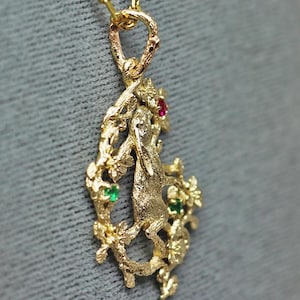 May include: A gold-toned pendant necklace featuring a detailed design of a creature within a floral frame. The pendant is adorned with red and green gemstones. The necklace hangs on a delicate gold chain.